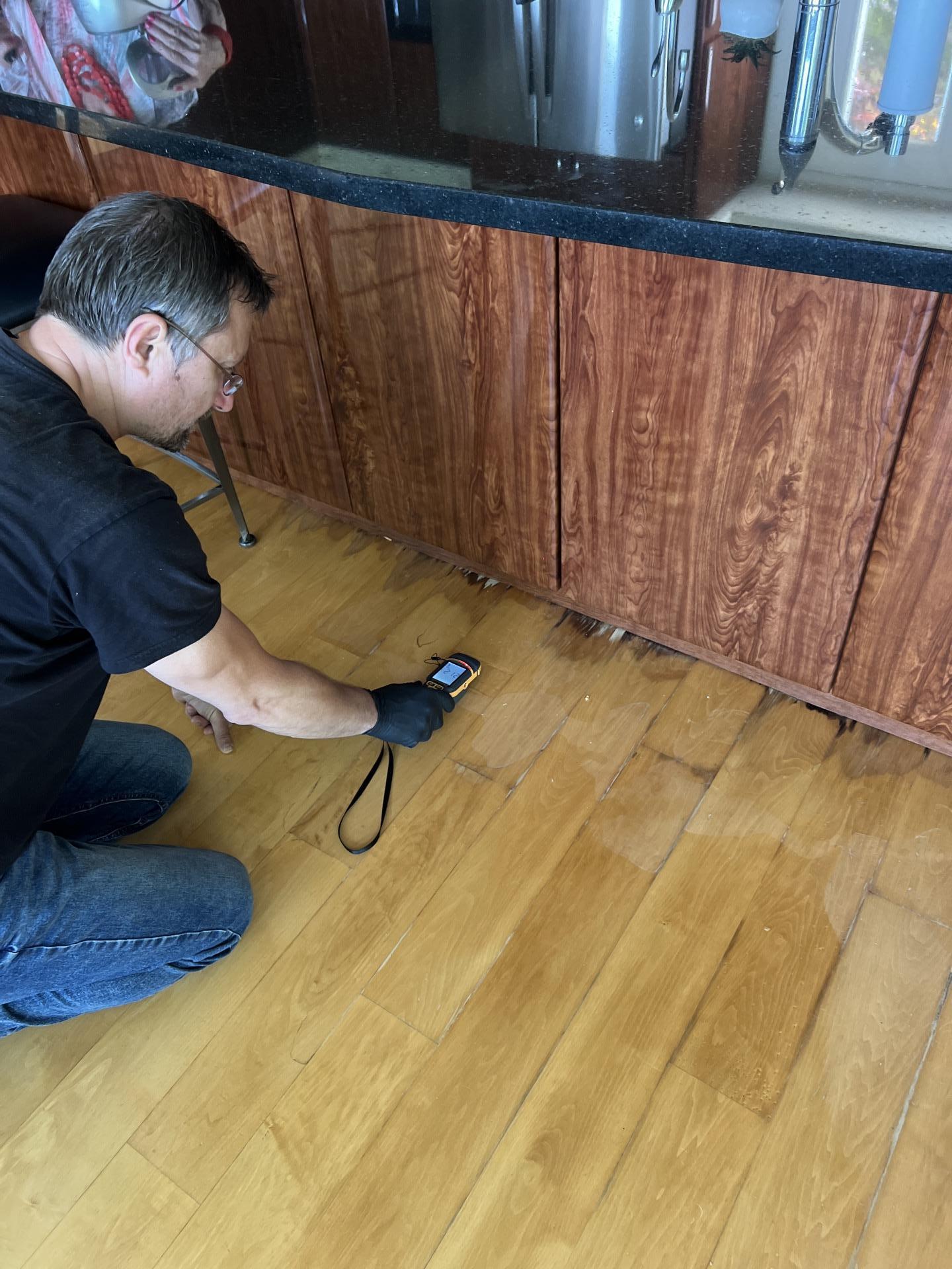 Technician measuring moisture levels in hardwood flooring near kitchen cabinetry during water mitigation